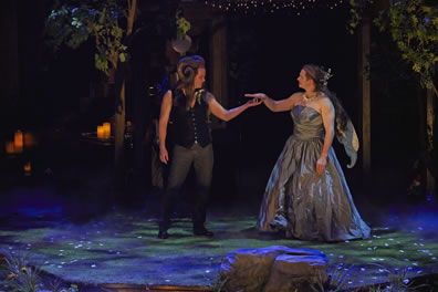 Oberon in black vest and jeans, no shirt, and with rams hors curling over the side of his head holds hands with Titania in silver, strapless ball gown, wings on the back, the stage floor dappled in blue and green lights and reflections of a mirror ball. Trees in the background, large rock in the foreground